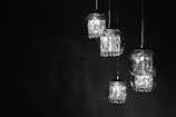 A series of glass pendant lights are suspended against a dark background. Each light contains multiple crystal-like elements, which catch and reflect the light, creating a luminous and elegant effect.