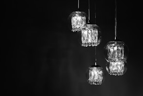 A series of glass pendant lights are suspended against a dark background. Each light contains multiple crystal-like elements, which catch and reflect the light, creating a luminous and elegant effect.