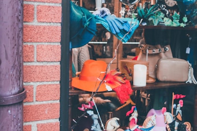 A store window display features a variety of items including a bright orange hat, a blue scarf, a beige handbag, and decorative elements like small figurines of horses with riders. There are also some green leafy decorations and a white candle placed among the items.