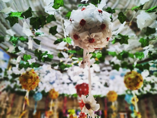 Colorful floral decorations hang from the ceiling, featuring white flowers with red centers surrounded by green leaves. The background includes a series of similar floral arrangements in various colors, adding depth and vibrancy to the scene.