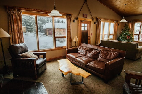 A cozy living room with leather sofas and a rustic wooden coffee table. Large windows allow natural light to illuminate the space, highlighting the snowy landscape visible outside. A decorated Christmas tree adds a festive touch to the room. Warm lighting fixtures and wooden accents contribute to the inviting atmosphere.