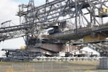 A massive industrial machine, likely a bucket-wheel excavator or conveyor system, constructed from steel beams and cables. It features a complex structure with various platforms, railings, and a large conveyor belt extending across the top. The components are painted in gray and beige tones, with some yellow highlights. The surrounding area includes a fence and sparse vegetation.