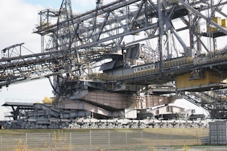 A massive industrial machine, likely a bucket-wheel excavator or conveyor system, constructed from steel beams and cables. It features a complex structure with various platforms, railings, and a large conveyor belt extending across the top. The components are painted in gray and beige tones, with some yellow highlights. The surrounding area includes a fence and sparse vegetation.
