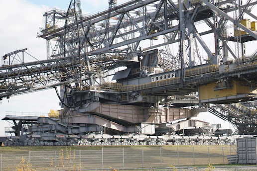 A massive industrial machine, likely a bucket-wheel excavator or conveyor system, constructed from steel beams and cables. It features a complex structure with various platforms, railings, and a large conveyor belt extending across the top. The components are painted in gray and beige tones, with some yellow highlights. The surrounding area includes a fence and sparse vegetation.