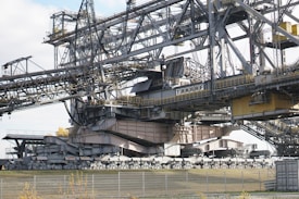 A massive industrial machine, likely a bucket-wheel excavator or conveyor system, constructed from steel beams and cables. It features a complex structure with various platforms, railings, and a large conveyor belt extending across the top. The components are painted in gray and beige tones, with some yellow highlights. The surrounding area includes a fence and sparse vegetation.