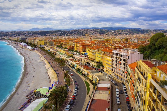 Aerial view of a vibrant coastal city featuring a long, sandy beach bordered by turquoise waters. Lined along the waterfront are tall yellow and orange buildings with red-tiled roofs. A curving promenade with palm trees runs parallel to the shoreline, accommodating people walking and cars. In the background, rolling hills with dense urban development extend into the distance under a partly cloudy sky.
