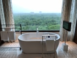 A modern bathroom features a freestanding white bathtub positioned near a large window with a scenic view of a lush green forest. Elegant patterned curtains frame the window, and a towel rack with neatly folded towels sits on the left. On the right, a mounted television adds a touch of luxury, and geometric patterns on the floor tiles enhance the contemporary design.