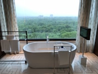 A modern bathroom features a freestanding white bathtub positioned near a large window with a scenic view of a lush green forest. Elegant patterned curtains frame the window, and a towel rack with neatly folded towels sits on the left. On the right, a mounted television adds a touch of luxury, and geometric patterns on the floor tiles enhance the contemporary design.