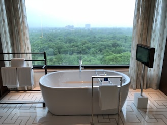 A modern bathroom features a freestanding white bathtub positioned near a large window with a scenic view of a lush green forest. Elegant patterned curtains frame the window, and a towel rack with neatly folded towels sits on the left. On the right, a mounted television adds a touch of luxury, and geometric patterns on the floor tiles enhance the contemporary design.