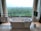 A modern bathroom features a freestanding white bathtub positioned near a large window with a scenic view of a lush green forest. Elegant patterned curtains frame the window, and a towel rack with neatly folded towels sits on the left. On the right, a mounted television adds a touch of luxury, and geometric patterns on the floor tiles enhance the contemporary design.