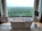 A modern bathroom features a freestanding white bathtub positioned near a large window with a scenic view of a lush green forest. Elegant patterned curtains frame the window, and a towel rack with neatly folded towels sits on the left. On the right, a mounted television adds a touch of luxury, and geometric patterns on the floor tiles enhance the contemporary design.