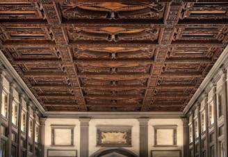 An ornate, richly detailed wooden ceiling with intricate carvings and decorative patterns dominates the view. The walls are lined with classical architectural elements, featuring columns and moldings in a neutral color palette.