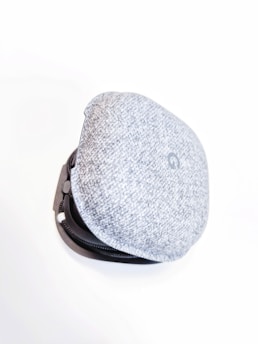 A soft, fabric-covered case with a rounded, cushion-like appearance. It is light grey with a subtle textured pattern and has a black zipper outlining the edge.