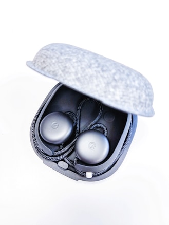 A pair of black earphones rests inside an open gray fabric case. The earphones have a circular design with a cord looped around them.