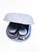 A pair of black wireless earbuds resting in a partially open gray fabric charging case. The earbuds have a sleek design with a slight shine and are neatly arranged in the case.
