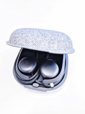 A pair of black wireless earbuds resting in a partially open gray fabric charging case. The earbuds have a sleek design with a slight shine and are neatly arranged in the case.