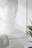 A photo with a minimalist aesthetic features a blurred and softly lit portrait of a person with light hair, partially obscured and rendered in pale tones. In the foreground, a white, modern chair is visible, and there is a green plant with long leaves in a white pot on the right side.