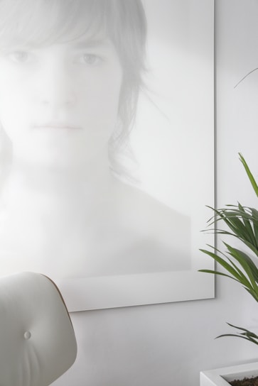 A photo with a minimalist aesthetic features a blurred and softly lit portrait of a person with light hair, partially obscured and rendered in pale tones. In the foreground, a white, modern chair is visible, and there is a green plant with long leaves in a white pot on the right side.