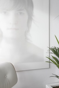 A photo with a minimalist aesthetic features a blurred and softly lit portrait of a person with light hair, partially obscured and rendered in pale tones. In the foreground, a white, modern chair is visible, and there is a green plant with long leaves in a white pot on the right side.
