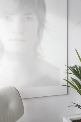 A photo with a minimalist aesthetic features a blurred and softly lit portrait of a person with light hair, partially obscured and rendered in pale tones. In the foreground, a white, modern chair is visible, and there is a green plant with long leaves in a white pot on the right side.