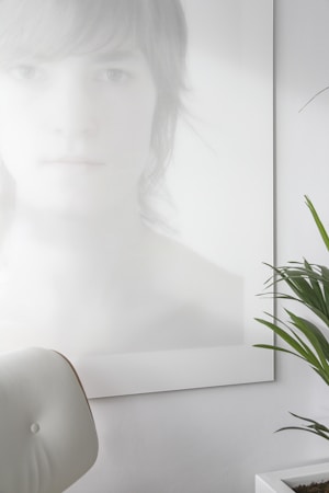 A photo with a minimalist aesthetic features a blurred and softly lit portrait of a person with light hair, partially obscured and rendered in pale tones. In the foreground, a white, modern chair is visible, and there is a green plant with long leaves in a white pot on the right side.