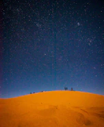 Wide shot of a group camping under a star-filled desert sky near towering sand dunes.