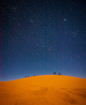 Wide shot of a group camping under a star-filled desert sky near towering sand dunes.