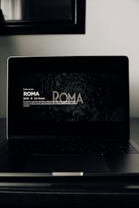 A dark-themed image featuring a laptop on a table. The screen displays text about the movie 'Roma', including details such as the year of release, duration, and a brief synopsis in Spanish. The surrounding environment appears dimly lit, with a mirror or framed artwork visible above the laptop.