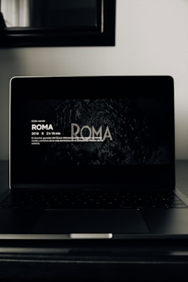A dark-themed image featuring a laptop on a table. The screen displays text about the movie 'Roma', including details such as the year of release, duration, and a brief synopsis in Spanish. The surrounding environment appears dimly lit, with a mirror or framed artwork visible above the laptop.