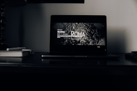 A dark room with a laptop open on a desk. The laptop screen displays a black and white scene from the film 'ROMA', with text about its duration and director. Beside the laptop, there are two closed notebooks and a pen.