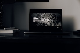 A dark room with a laptop open on a desk. The laptop screen displays a black and white scene from the film 'ROMA', with text about its duration and director. Beside the laptop, there are two closed notebooks and a pen.