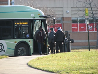A public bus with the route sign '8 to Telegraph' is stopped on a sidewalk. Several individuals are near the bus, with some entering and others standing close by. In the background, there is a commercial building with red signage, and a small patch of green grass is visible in the foreground.