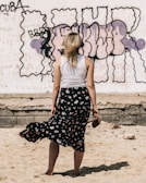 A woman with blonde hair stands barefoot in the sand, holding her shoes in one hand. She is wearing a white sleeveless top and a long, dark floral skirt. The background features a graffiti-covered wall with various colors and text, including bold, dark outlines.