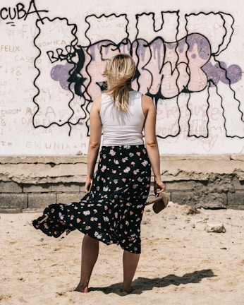 A woman with blonde hair stands barefoot in the sand, holding her shoes in one hand. She is wearing a white sleeveless top and a long, dark floral skirt. The background features a graffiti-covered wall with various colors and text, including bold, dark outlines.