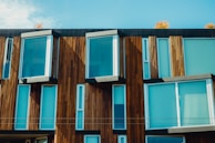 A modern building facade featuring large glass windows and wooden paneling. The structure’s design includes protruding rectangular sections, creating a dynamic interplay of light and shadow. Autumnal trees are partially visible at the top, adding a touch of nature.
