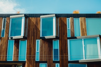 A modern building facade featuring large glass windows and wooden paneling. The structure’s design includes protruding rectangular sections, creating a dynamic interplay of light and shadow. Autumnal trees are partially visible at the top, adding a touch of nature.