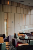 A modern café interior with round tables and multi-colored chairs. The space has wooden paneling on the walls and a soft lighting ambience. Small decorative cakes are placed on the tables.