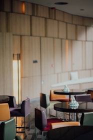 A modern café interior with round tables and multi-colored chairs. The space has wooden paneling on the walls and a soft lighting ambience. Small decorative cakes are placed on the tables.