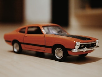 A small, detailed model car with an orange body and a black stripe on the hood is displayed on a wooden surface. The car features silver details on the front, including the grille and bumper.