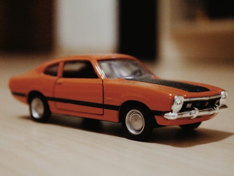 A small, detailed model car with an orange body and a black stripe on the hood is displayed on a wooden surface. The car features silver details on the front, including the grille and bumper.