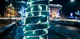 A tree is wrapped with glowing white string lights, set against a nighttime cityscape with various buildings and blurred colorful lights in the background. The area is lightly dusted with snow, hinting at a festive or winter setting.