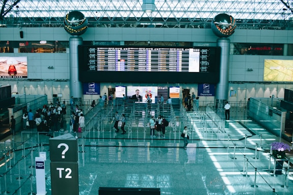 A busy airport terminal features a large digital departure board displaying flight information. Several small groups of travelers can be seen in various parts of the terminal, some clustered near a queue. The area is illuminated by natural light filtering through the glass ceiling. Two large metallic spheres are mounted near the board as decorative elements. Signage indicates this is Terminal 2.