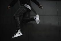 A person is captured mid-jump, wearing stylish Nike sneakers that stand out against their dark clothing and the dark background. The angle of the shot highlights the shoes and the dynamic pose of the person.