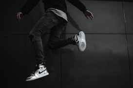A person is captured mid-jump, wearing stylish Nike sneakers that stand out against their dark clothing and the dark background. The angle of the shot highlights the shoes and the dynamic pose of the person.