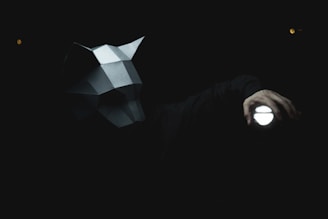 A person wearing a geometric wolf-shaped mask is holding a flashlight in a dark environment. The light from the flashlight casts a prominent glow, illuminating parts of the mask and the hand holding the flashlight.