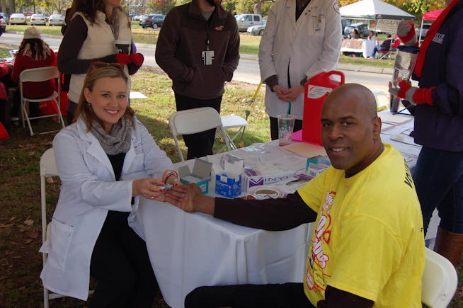 A man is receiving a medical check-up or test from a female healthcare professional who is sitting at a table outside. The man is smiling and wearing a yellow t-shirt, while the professional is dressed in a white coat. There are several people standing around, likely part of an event or fair. Medical supplies and informational materials are scattered on the table.
