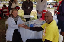 A man is receiving a medical check-up or test from a female healthcare professional who is sitting at a table outside. The man is smiling and wearing a yellow t-shirt, while the professional is dressed in a white coat. There are several people standing around, likely part of an event or fair. Medical supplies and informational materials are scattered on the table.