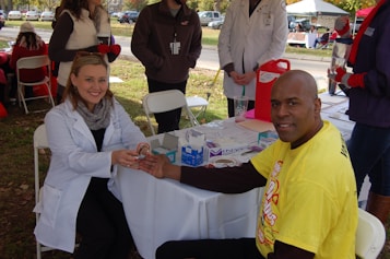 A man is receiving a medical check-up or test from a female healthcare professional who is sitting at a table outside. The man is smiling and wearing a yellow t-shirt, while the professional is dressed in a white coat. There are several people standing around, likely part of an event or fair. Medical supplies and informational materials are scattered on the table.