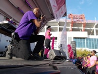 A photographer wearing a purple shirt kneels on stage taking photos at an outdoor event. A person dressed in pink, possibly a performer or speaker, stands on stage holding a pink ribbon, while a crowd of people in pink attire watches. Balloons and stage equipment are visible, with a large building and clear blue sky in the background.
