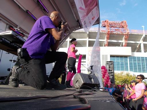 A photographer wearing a purple shirt kneels on stage taking photos at an outdoor event. A person dressed in pink, possibly a performer or speaker, stands on stage holding a pink ribbon, while a crowd of people in pink attire watches. Balloons and stage equipment are visible, with a large building and clear blue sky in the background.